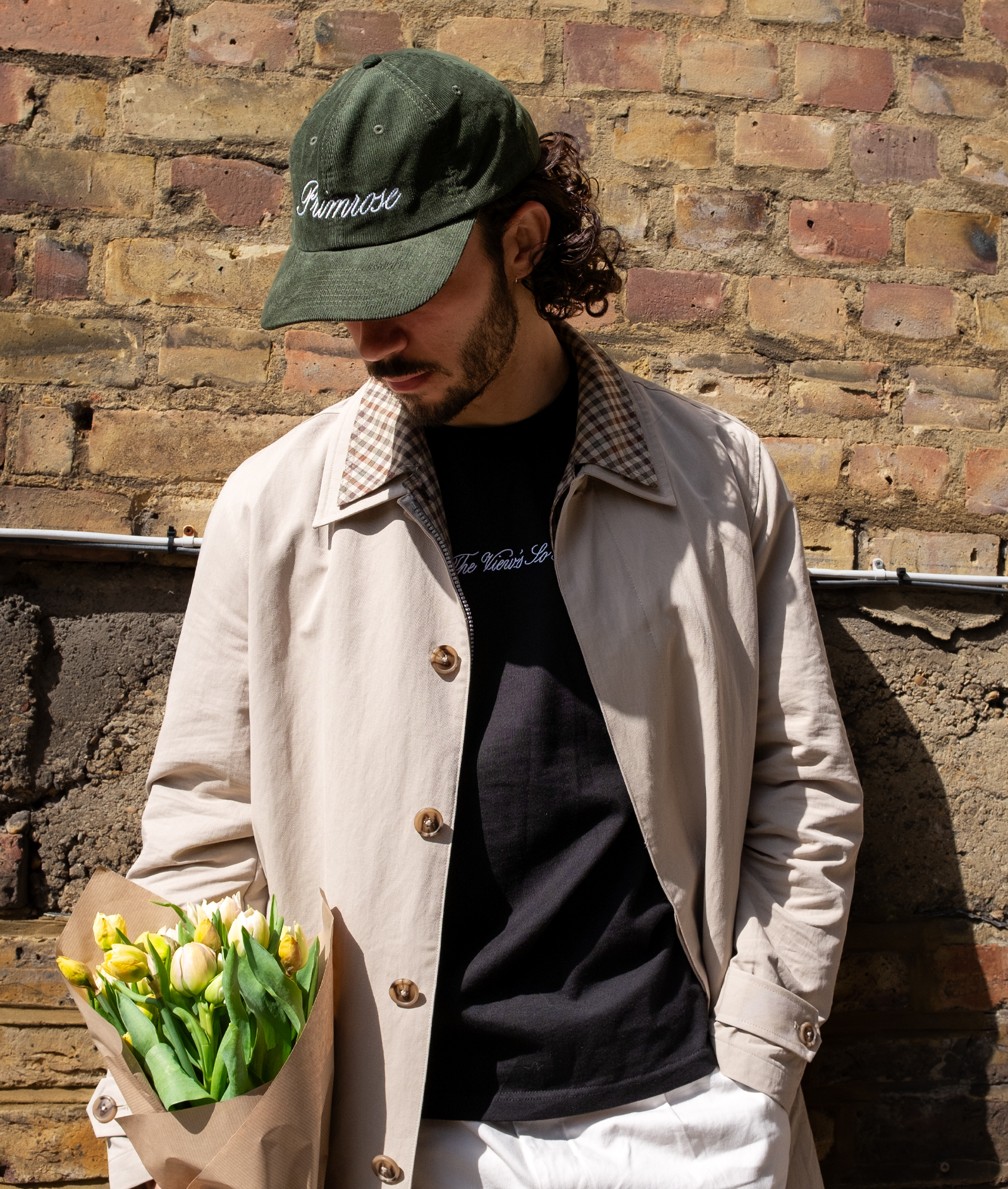Man holding flowers against a brick wall, wearing Primrose London embroidered cap.