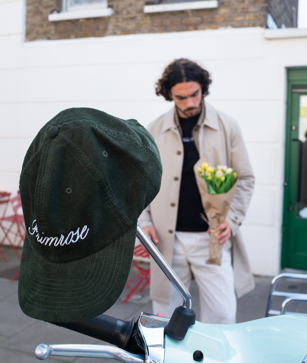 Green cap with 'Primrose' branding on a scooter, man holding flowers in the background.