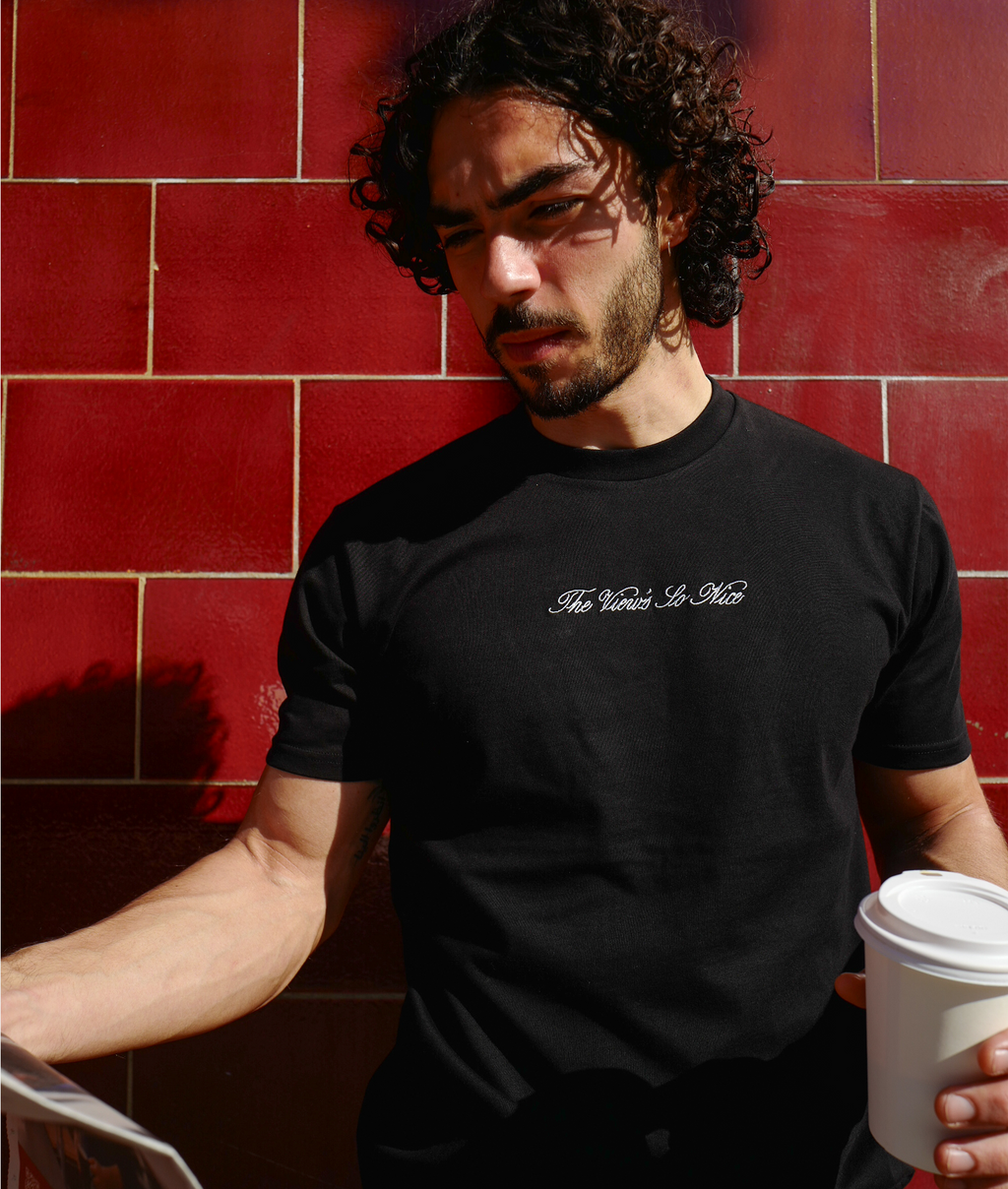 Man wearing a black t-shirt with text, holding a white cup against a red tiled wall.