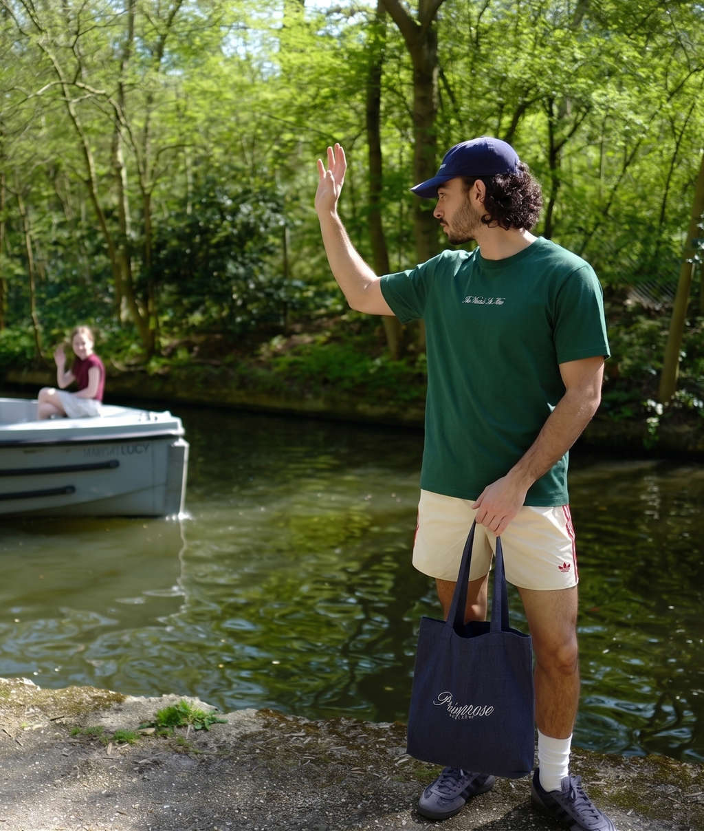 Man waving at someone on a boat by a lake with trees in the background, wearing a Primrose London tee