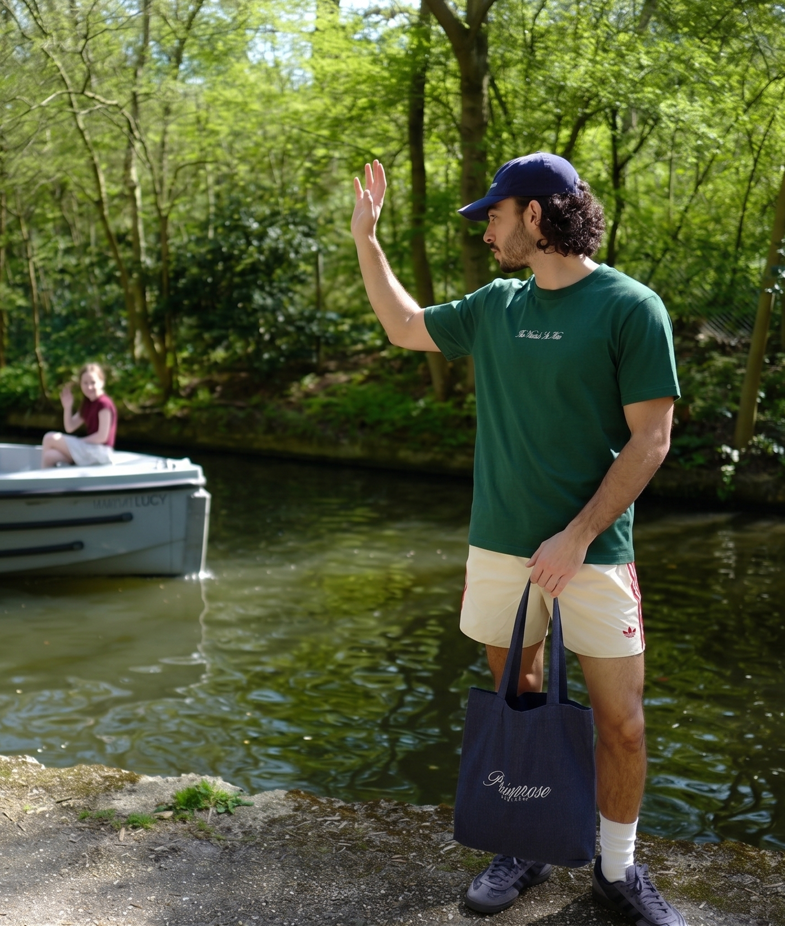 Man waving at someone on a boat by a lake with trees in the background, wearing a Primrose London tee